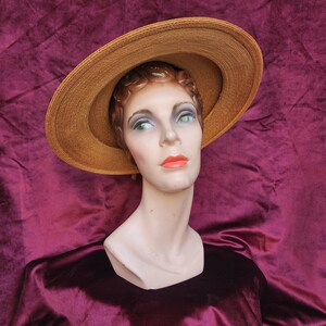 May include: A brown wide-brimmed straw hat with a slightly curled brim. The hat is worn by a mannequin with a neutral expression. The mannequin is wearing a burgundy velvet dress.