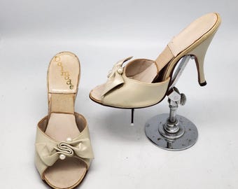 Puede incluir: Un par de zapatillas de satén blanco roto con tacón, detalle de lazo y adornos de perlas. Las zapatillas están etiquetadas con "Dolmode" en la plantilla.