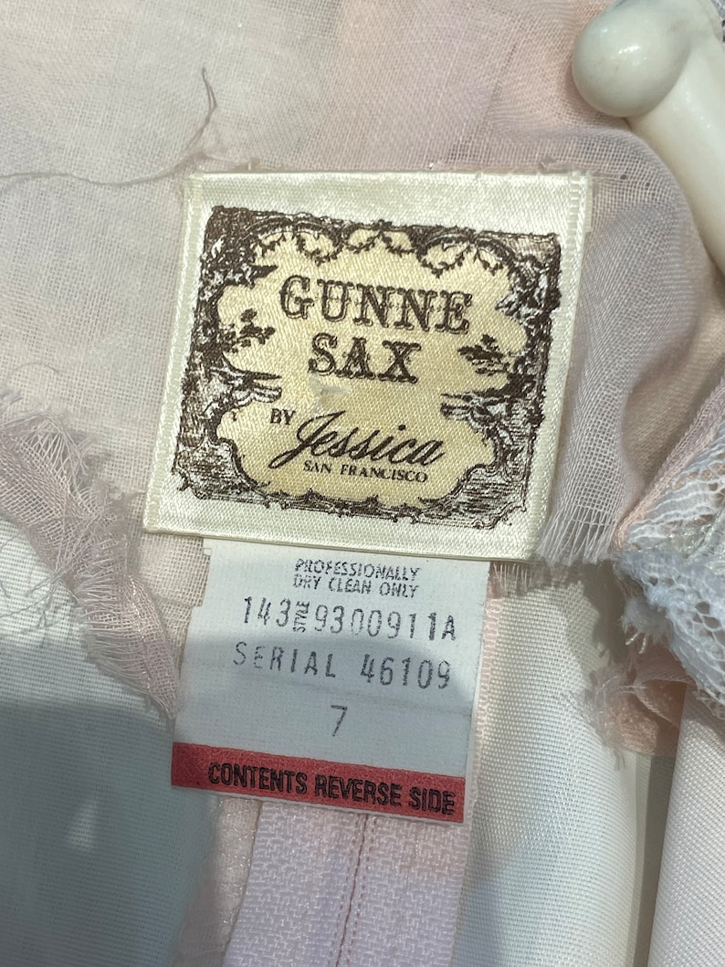 May include: A white tag with brown text that reads "GUNNE SAX by Jessica SAN FRANCISCO". Below the text is a smaller white tag with brown text that reads "PROFESSIONALLY DRY CLEAN ONLY 1439300911A SERIAL 46109 7 CONTENTS REVERSE SIDE".