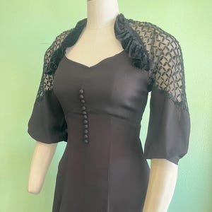 May include: A black vintage dress with a sweetheart neckline and a row of buttons down the front. The dress features short sleeves and is paired with a black lace bolero with a ruffled collar. The dress is displayed on a mannequin.