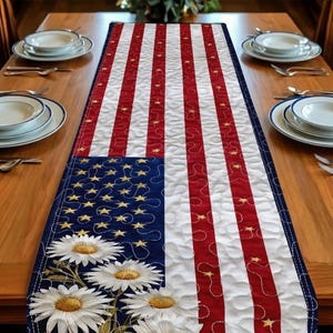 May include: A patriotic table runner featuring the American flag design. The runner has red and white stripes, a blue field with gold stars, and white daisies. The runner is placed on a wooden table, ready for a festive occasion.