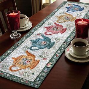 Könnte beinhalten: Gesteppter Tischläufer mit bunten Teekannen-Designs. Der Läufer hat einen weißen Hintergrund mit floralen Akzenten und einem grünen Rand. Teekannen in Blau, Gelb, Rosa und Orange. Zwei rote Kerzenhalter und Teetassen auf dem Tisch.