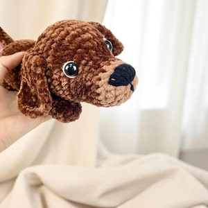 K&ouml;nnte beinhalten: Ein brauner geh&auml;kelter Hund mit schwarzer Nase und gro&szlig;en, runden schwarzen Augen. Das Pl&uuml;schtier ist aus einem weichen, strukturierten Garn gefertigt und hat Schlappohren. Der Hund ist ein handgefertigter Artikel.