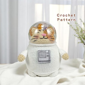May include: White crocheted astronaut with a cat face inside a clear plastic helmet. The astronaut has a black and white embroidered patch on the chest. Crochet Pattern.