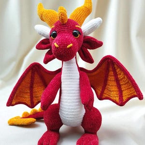 May include: A handmade crochet dragon in shades of red, orange, yellow, and white. The dragon has large, expressive eyes, horns, wings, and a tail. The wings are orange with red outlines. The dragon is a whimsical, soft toy.