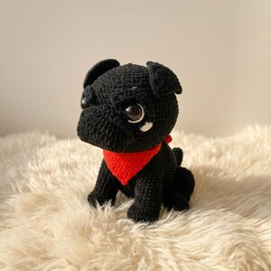 May include: A handmade black crochet pug dog with large, expressive eyes and a bright red bandana. The plush toy is sitting on a fluffy, cream-colored surface. The pug's features are detailed, showcasing the craftsmanship of the crochet work.