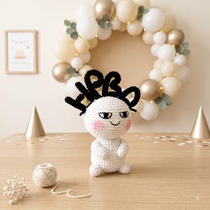 May include: A white crocheted doll with a round head, pink cheeks, and black embroidered eyes and mouth. The doll features a black crocheted topper with the letters HPBD in a stylized font. The doll has a small body with short arms and legs.