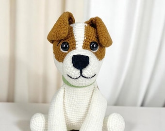 Puede incluir: Un juguete de perro de ganchillo hecho a mano en blanco, marrón y negro. El perro tiene orejas marrones, una cabeza blanca y marrón y una nariz negra. Lleva un collar verde y está sentado sobre una superficie blanca.