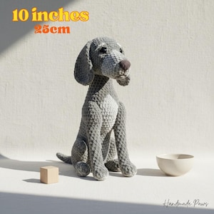 Könnte beinhalten: Ein handgefertigter, grauer Häkelhund, etwa 25 cm hoch, mit weißer Brust und brauner Nase. Der Hund sitzt, mit einem kleinen Holzblock und einer cremefarbenen Schüssel in der Nähe. Der Text "Handmade Paws" ist sichtbar.
