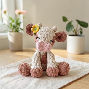 May include: A handmade crochet cow plushie in shades of white, pink, and brown, with a yellow flower on its head. The cow is sitting on a white cloth, with its eyes closed. The plushie is a soft, cuddly toy.