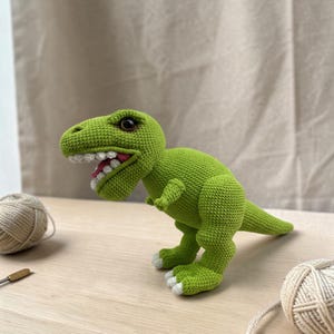 May include: A green crocheted Tyrannosaurus Rex dinosaur toy with white teeth and a red tongue. The dinosaur has white accents on its feet and dark eyes. The toy is posed on a wooden surface.