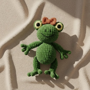 May include: A green crocheted frog plush toy with large, round eyes and a small orange bow on its head. The frog has outstretched arms and legs, and a smiling face. The plush is made of a soft, textured yarn.