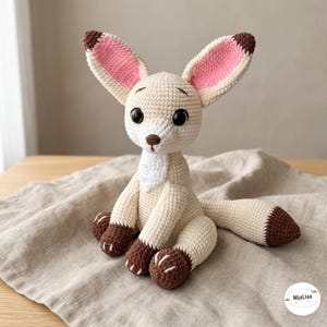 May include: A crocheted fennec fox plush toy with large ears featuring pink inner sections, a cream-colored body, dark brown paws, and a dark brown tail tip. The toy has black plastic eyes and a small brown embroidered nose. Text MjaLisa is visible.