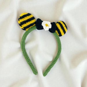 May include: A handmade crochet headband featuring yellow and black striped bee antennae and a black bow with a white flower accent. The headband is green and sits on a white surface.