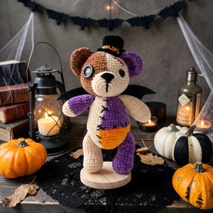May include: A crocheted teddy bear with a patchwork design in brown, cream, purple, and orange yarn. The bear features a black top hat, bat wings, a button eye, and stitched details. It stands on a round wooden base, showcasing a multi-colored plush texture.