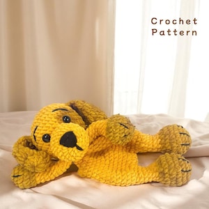 Puede incluir: Un juguete de perro de ganchillo amarillo hecho a mano con ojos y nariz negros, acostado sobre una superficie blanca. El perro tiene un aspecto texturizado y afelpado. El texto "Crochet Pattern" es visible.
