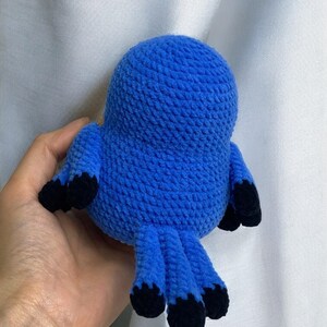 Puede incluir: Un juguete de p&aacute;jaro de crochet azul con pies de crochet negro. El p&aacute;jaro est&aacute; sentado en una mano.