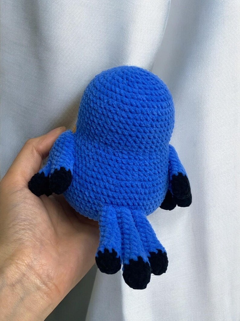 Puede incluir: Un juguete de p&aacute;jaro de crochet azul con pies de crochet negro. El p&aacute;jaro est&aacute; sentado en una mano.