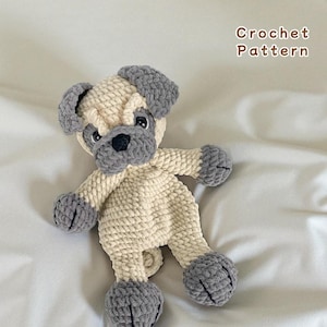 Könnte beinhalten: Ein handgehäkelter Mops-Hund in Creme und Grau. Der Hund hat graue Ohren, Pfoten und eine Schnauze. Der Text "Crochet Pattern" befindet sich in der oberen rechten Ecke. Der Mops liegt auf einer weißen Oberfläche.