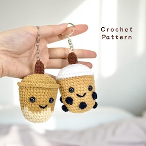 Puede incluir: Dos amuletos de té de burbujas de ganchillo. Uno es marrón con espuma de leche blanca y el otro es blanco con té de leche marrón y perlas de boba negras. Ambos tienen caras sonrientes con ojos negros.