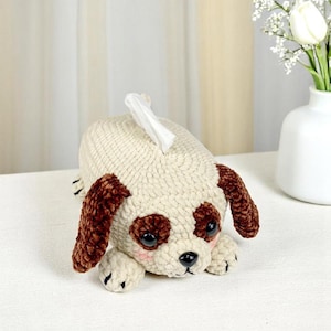 Może przedstawiać: Tkany na szydełku pokrowiec na pudełko chusteczek w kształcie psa z brązowymi uszami i białym ciałem. Pies ma czarne oczy i czarny nos.