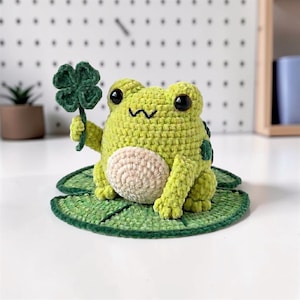 May include: A crocheted light green frog with a cream-colored belly, black plastic eyes, and a stitched mouth. The frog is sitting on a flat, circular green lily pad and is holding a small, dark green four-leaf clover in its right hand.