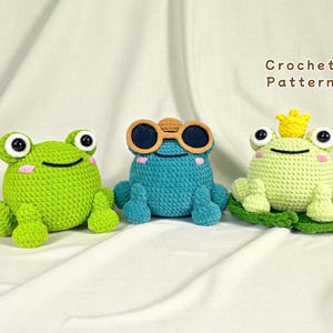 May include: Three crocheted frog toys in shades of green and blue. One frog wears sunglasses, and another wears a crown. The text "Crochet Pattern" is visible in the background. These handmade toys are perfect for children.