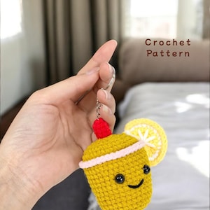 Puede incluir: Un llavero de ganchillo amarillo hecho a mano con forma de cóctel, con una rodaja de limón y una cereza roja. El llavero tiene una cara sonriente y el texto "Crochet Pattern".