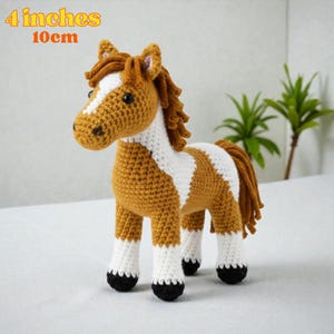 Puede incluir: Un juguete de caballo hecho a mano con ganchillo, de aproximadamente 10 cm de altura. El caballo presenta una combinación de colores marrón y blanco con una melena y cola detalladas. El texto "4 inches 10cm" está en la esquina superior izquierda.