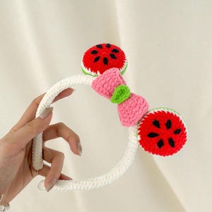 May include: White crocheted headband featuring two watermelon slices and a pink bow. The watermelon slices are red with black seeds and green and white trim. The bow has a green leaf accent.