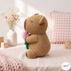 May include: A brown crocheted stuffed animal with a dark brown snout and closed eyes, holding a small pink flower with a green stem. The plush toy features a textured yarn construction and is positioned in profile, showcasing its rounded shape and ears.