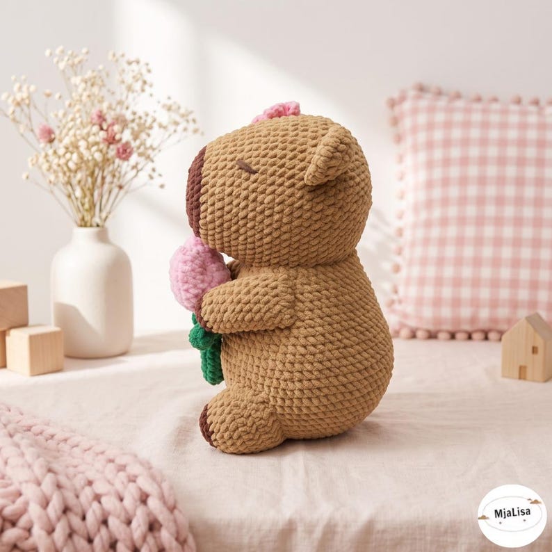 May include: A brown crocheted stuffed animal with a dark brown snout and closed eyes, holding a small pink flower with a green stem. The plush toy features a textured yarn construction and is positioned in profile, showcasing its rounded shape and ears.