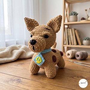 Op de afbeelding: Een gehaakte hond als knuffel in bruin met donkerbruine vlekken, puntige oren en zwarte plastic ogen. Het draagt een lichtblauwe halsband met een ruitvormig label waarop de gele letters SD staan afgebeeld in een zachte textuur.
