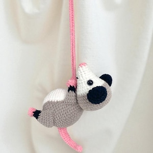 May include: A handmade crochet opossum toy in gray, white, and pink, hanging from a pink string. The opossum has dark blue ears and a black eye. The toy is suspended against a white background.