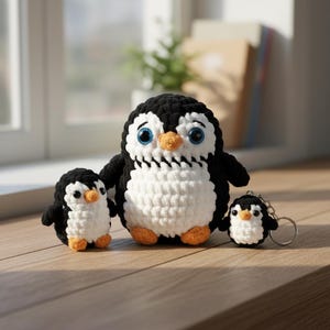May include: Three handmade crochet penguins in black, white, and orange. The largest penguin is in the centre, flanked by a smaller penguin on the left and a keychain penguin on the right. The penguins have blue eyes and orange beaks.