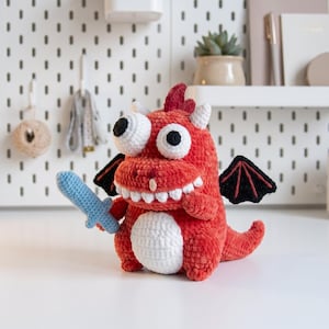 May include: A red crocheted dragon plush toy with large white eyes, a white belly, and black wings. The dragon holds a small light blue crocheted sword in its right hand and features a wide, toothy grin with white yarn teeth.