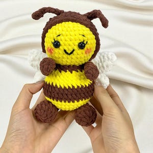 May include: A handmade crochet bee plushie. The bee features a yellow body with brown stripes, brown antennae, and small white wings. The bee has a smiling face with rosy cheeks and is held by two hands.