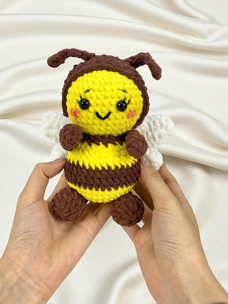 May include: A handmade crochet bee plushie. The bee features a yellow body with brown stripes, brown antennae, and small white wings. The bee has a smiling face with rosy cheeks and is held by two hands.