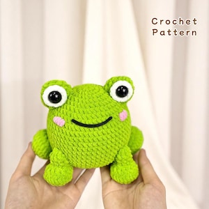Könnte beinhalten: Eine gehäkelte grüne Froschfigur mit großen weißen und schwarzen Augen, rosa Wangen und einem lächelnden Mund. Der Frosch ist handgefertigt und hat eine strukturierte Oberfläche. Der Text "Crochet Pattern" ist im Hintergrund zu sehen.