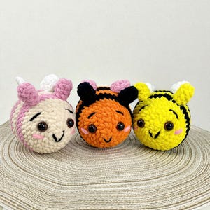 May include: Three crocheted bee toys in pink, orange, and yellow. Each bee has black eyes, a smiling mouth, and small wings. The pink bee has pink and white stripes. The orange bee has black stripes. The yellow bee has black stripes.