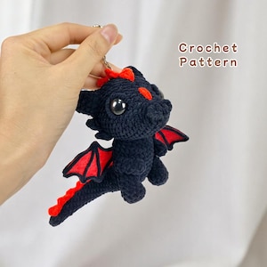 May include: A black and red crocheted dragon keyring. The dragon has a small, black body with red wings and a red tail. The dragon has black eyes and a red nose.