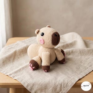 Peut inclure: Un animal en peluche capybara au crochet avec un corps beige clair, un museau marron foncé et des pieds marron foncé avec de petites touches roses. Le jouet a des yeux en plastique noir et une petite tache rose sur la joue. Le texte MjaLisa est visible.