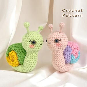 May include: Two crocheted snail toys. One is light green with a green and yellow shell. The other is pink with a shell of pink, purple, and blue. Both have black eyes, rosy cheeks, and antennae. The text "Crochet Pattern" is in the upper right.