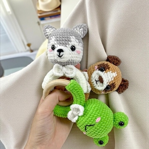 May include: Crocheted baby rattles in the shape of a gray and white husky, a brown and white bear, and a green frog. The husky rattle has a wooden ring and a white bow. The frog has a white flower and pink cheeks.