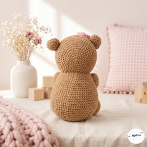 May include: A brown crochet teddy bear toy with a small pink flower detail on its head. The bear is crafted from thick yarn with a visible stitched texture, featuring rounded ears and a rounded body, shown from the back view in a neutral setting.
