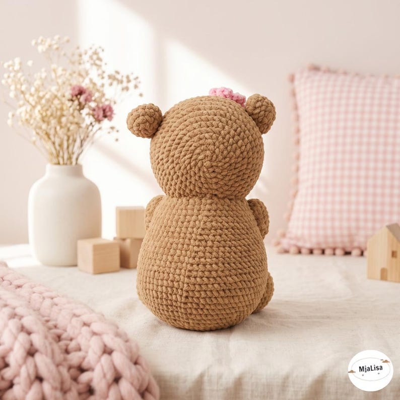May include: A brown crochet teddy bear toy with a small pink flower detail on its head. The bear is crafted from thick yarn with a visible stitched texture, featuring rounded ears and a rounded body, shown from the back view in a neutral setting.