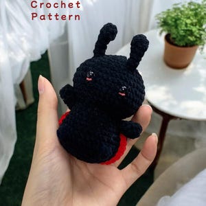 Puede incluir: Patrón de crochet para un juguete amigurumi de mariquita negro y rojo. La mariquita tiene una cara linda con ojos negros y mejillas rosadas.