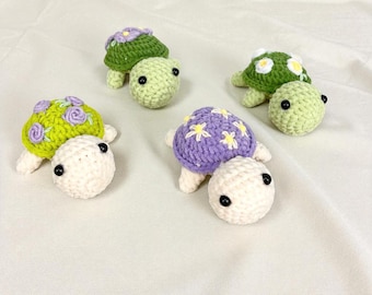May include: Six handmade crochet turtles in a variety of colours. The turtles have green, yellow, and purple shells with floral and star designs. Each turtle has a white head and small black eyes, creating a whimsical aesthetic.