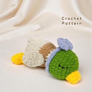 May include: A handmade crochet duck toy in a resting pose. The duck features a green head, yellow beak and feet, a white and brown body, and a blue accent. The words "Crochet Pattern" are in the background.