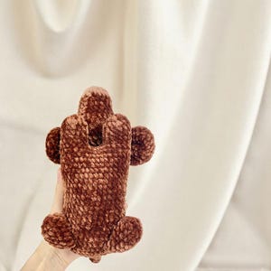 K&ouml;nnte beinhalten: Eine braune, pl&uuml;schige, geh&auml;kelte Handyh&uuml;lle. Die H&uuml;lle hat einen rechteckigen K&ouml;rper mit abgerundeten Kanten und kreisf&ouml;rmigen Akzenten. Die H&uuml;lle besteht aus einem weichen, strukturierten Material.